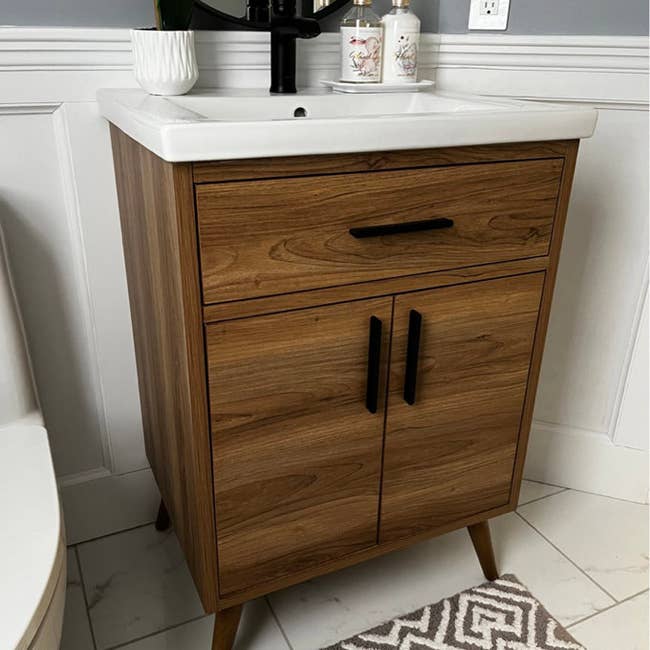 Wooden bathroom vanity with black handles, featuring a white basin, soap dispensers, and a patterned rug nearby