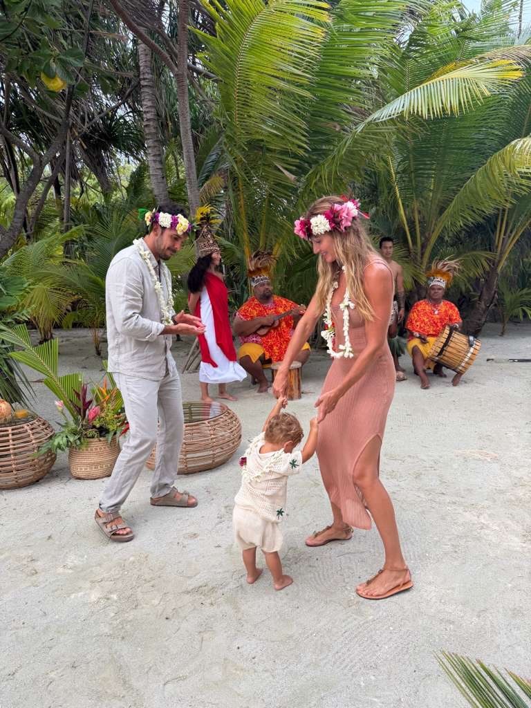 Gisele Bundchen celebrating her birthday with family on a beach, with tropical foliage and traditional musicians in the background.