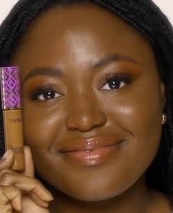 Person smiling while holding a tube of Tarte concealer beside their face, indicating a beauty product for shopping