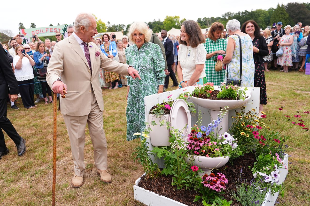 A man gestures towards a garden display featuring flowers planted in a toilet with people observing in an outdoor event setting