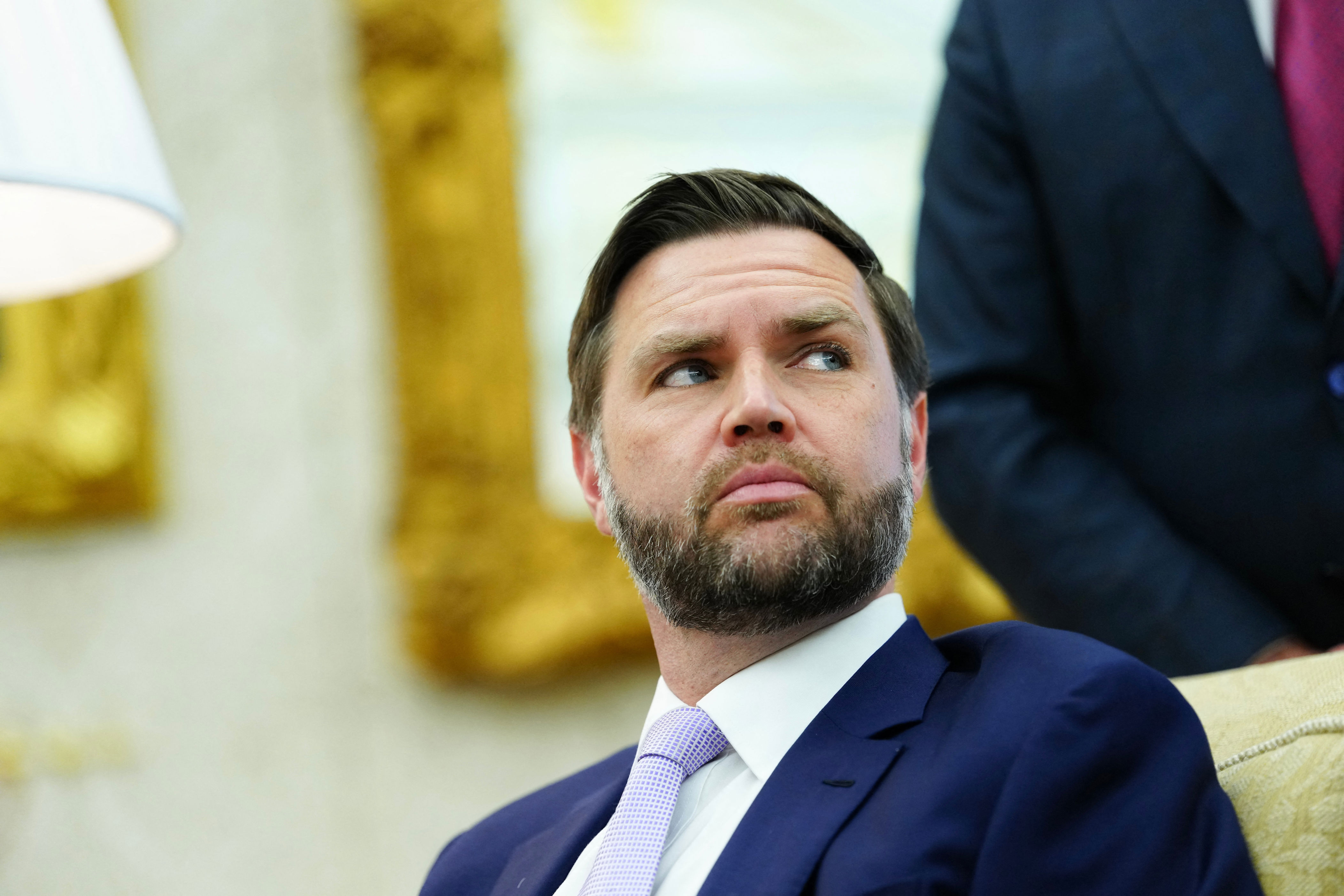 A man in a suit with a light tie sits in an official setting, looking to the side
