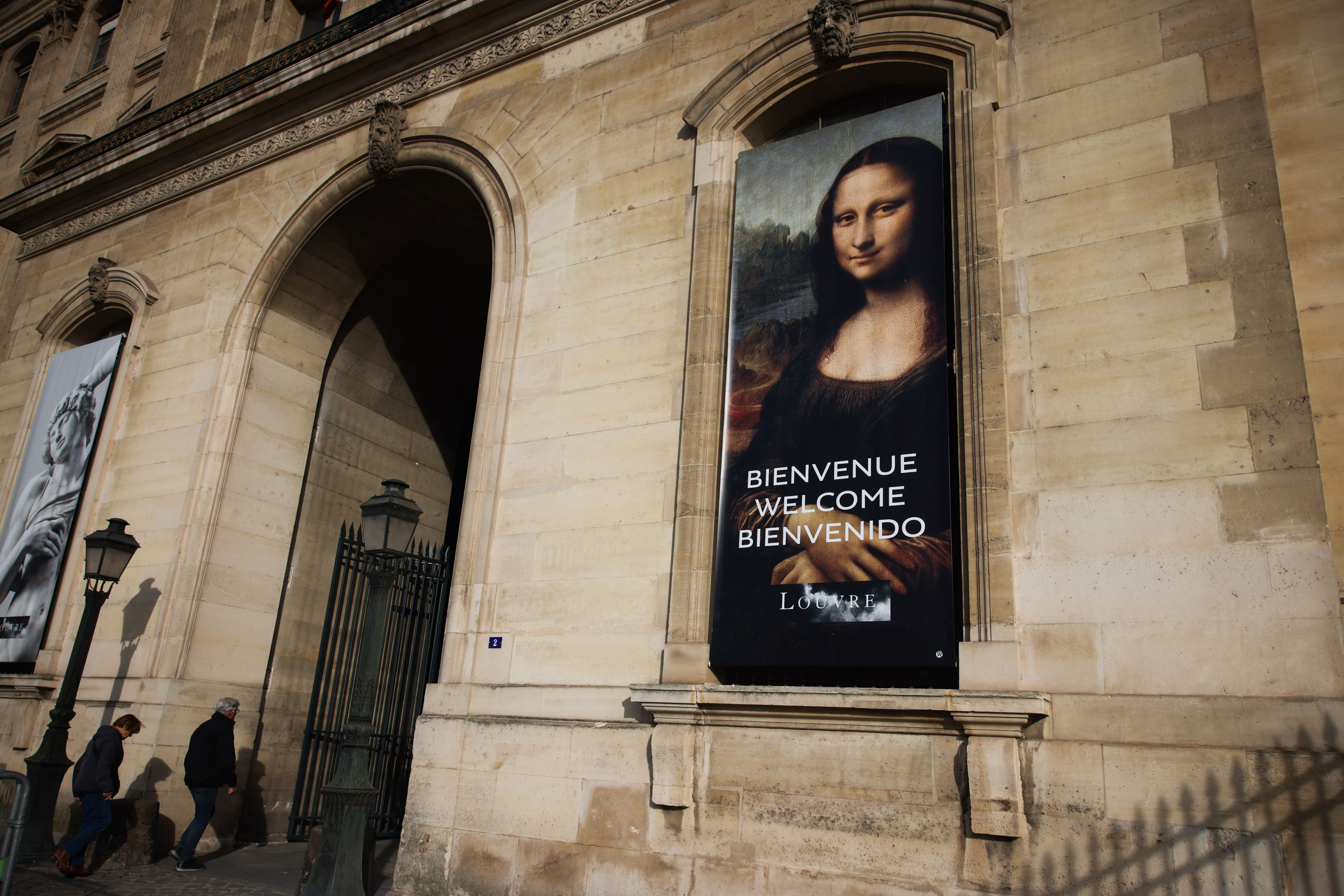 Banner at museum entrance showing the Mona Lisa with "Bienvenue, Welcome, Bienvenido" text. Visitors walk nearby