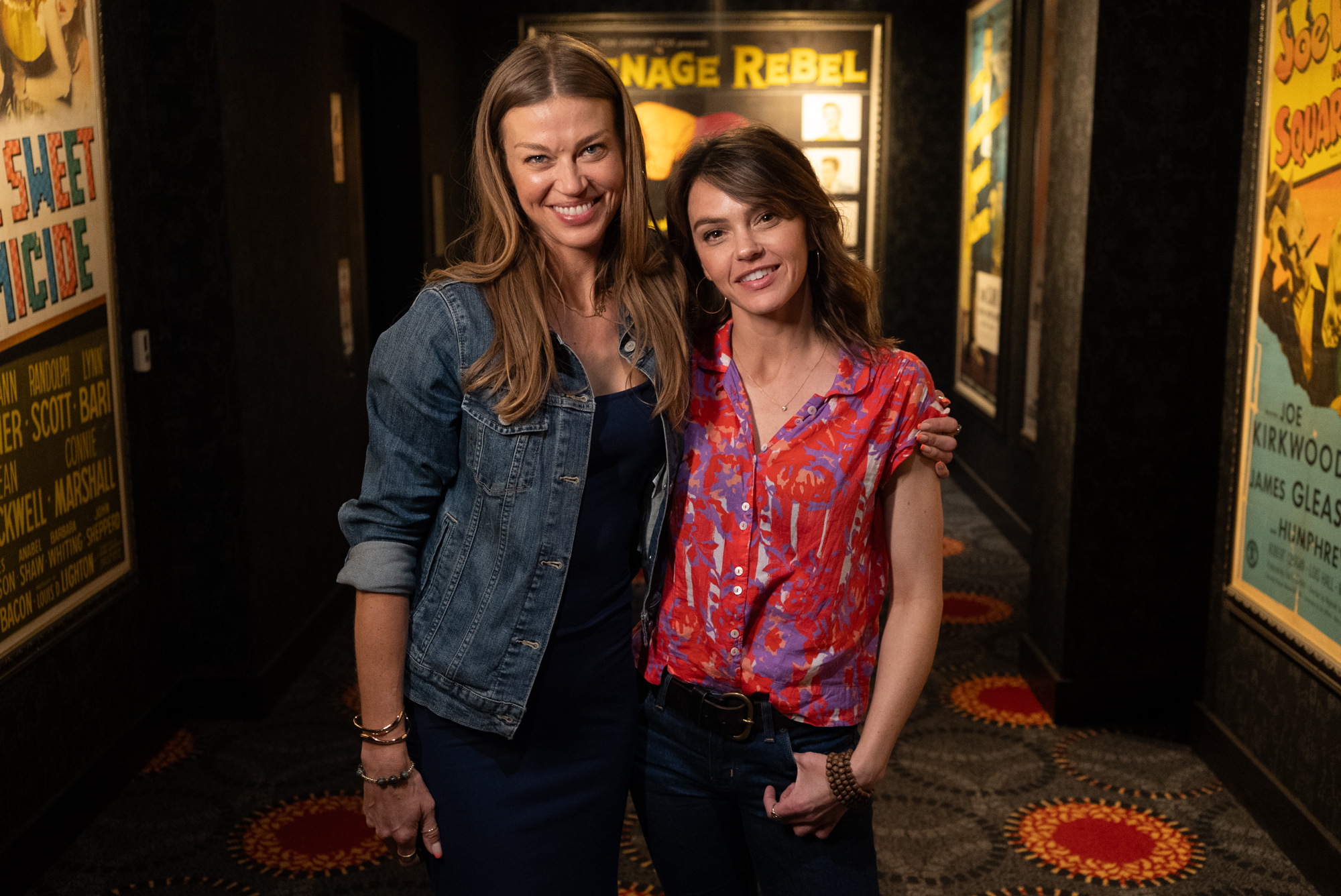 Adrianne Palicki and Aimee Teegarden smiling in a hallway with movie posters.