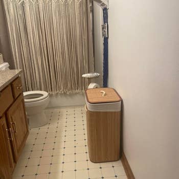 Bathroom with pattern tile floor, wooden cabinet, trash bin, and toilet. Beige macramé-style curtain hangs near the toilet