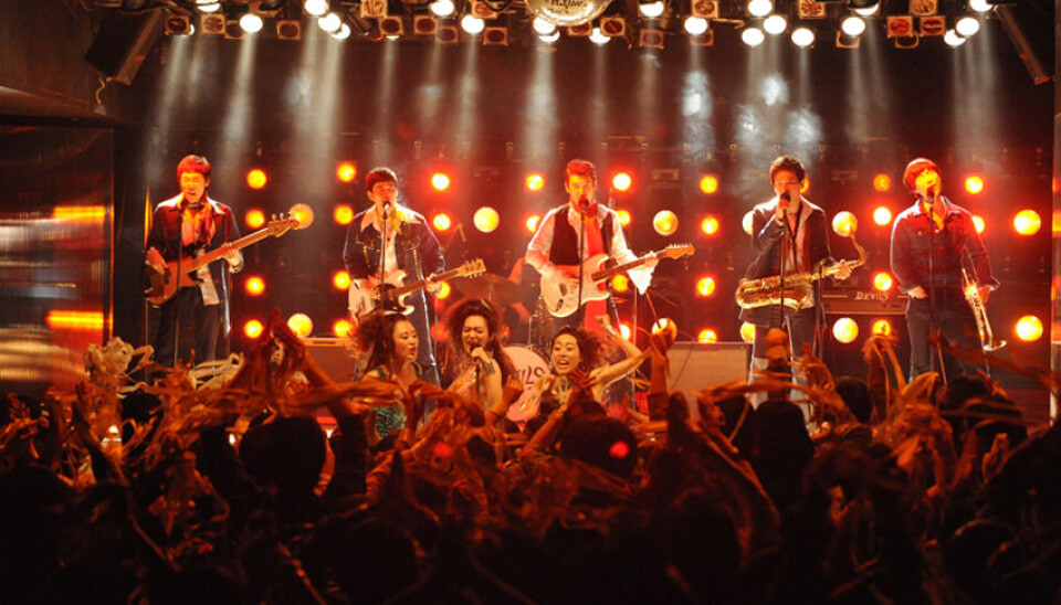 Rock band playing on stage before an audience under bright orange concert lighting.