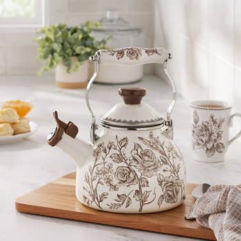 Vintage-style floral tea kettle with ornate handle on wooden board, matching mug nearby. Perfect for adding elegance to kitchen decor