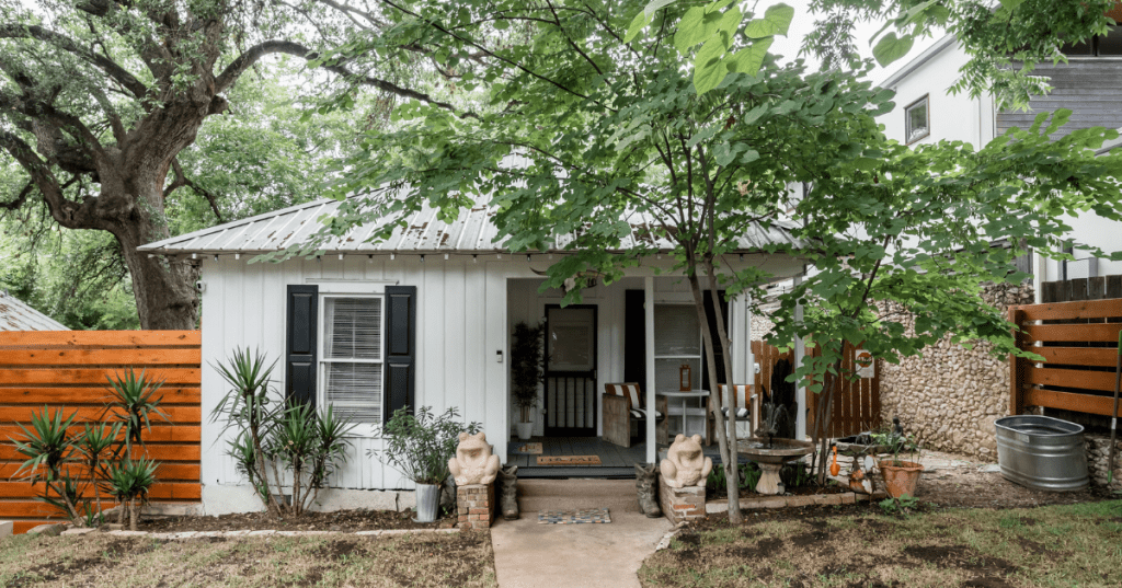 A charming white Craftsman bungalow Airbnb in Austin, Texas, featuring a small covered front porch, dark shutters, a brick walkway, and a wooden privacy fence surrounded by lush green trees.