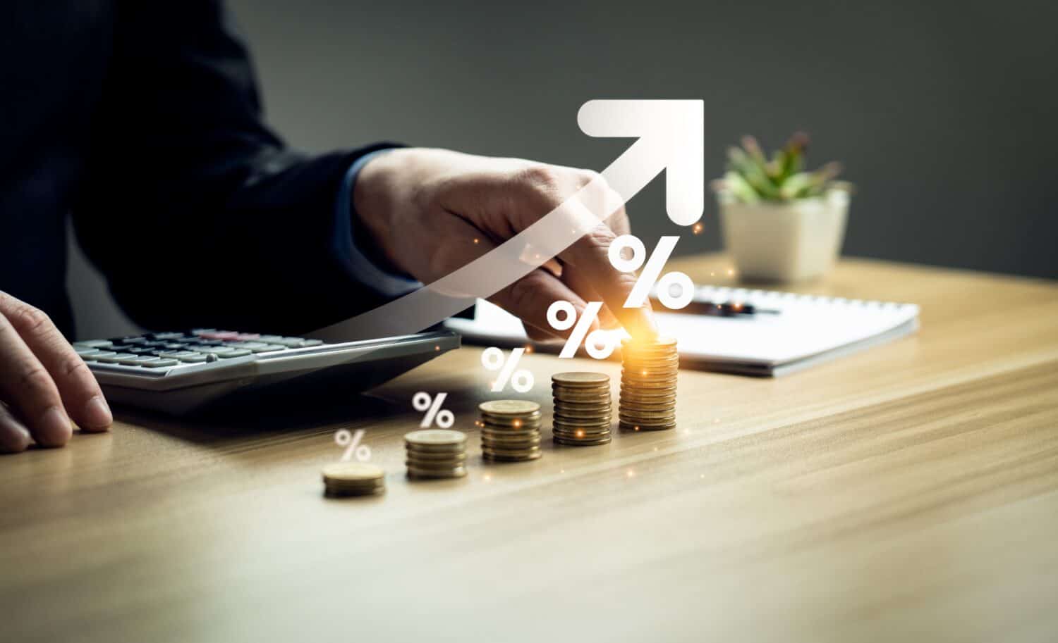 A person's hand in a business suit is positioned over a calculator on a light brown wooden desk. To the right, there are four progressively taller stacks of golden coins. White percentage symbols and a large white upward-curving arrow are overlaid on the scene, visually representing financial growth. In the background, a white notebook with a pen and a small green plant in a white pot are visible.