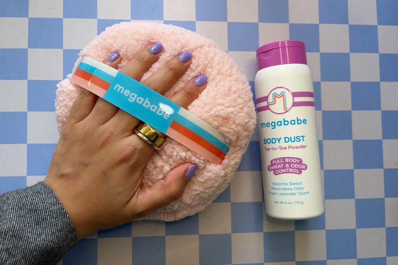 A hand with a ring and painted nails resting on a Megababe fluffy pad next to a Megababe Body Dust container on a tiled surface