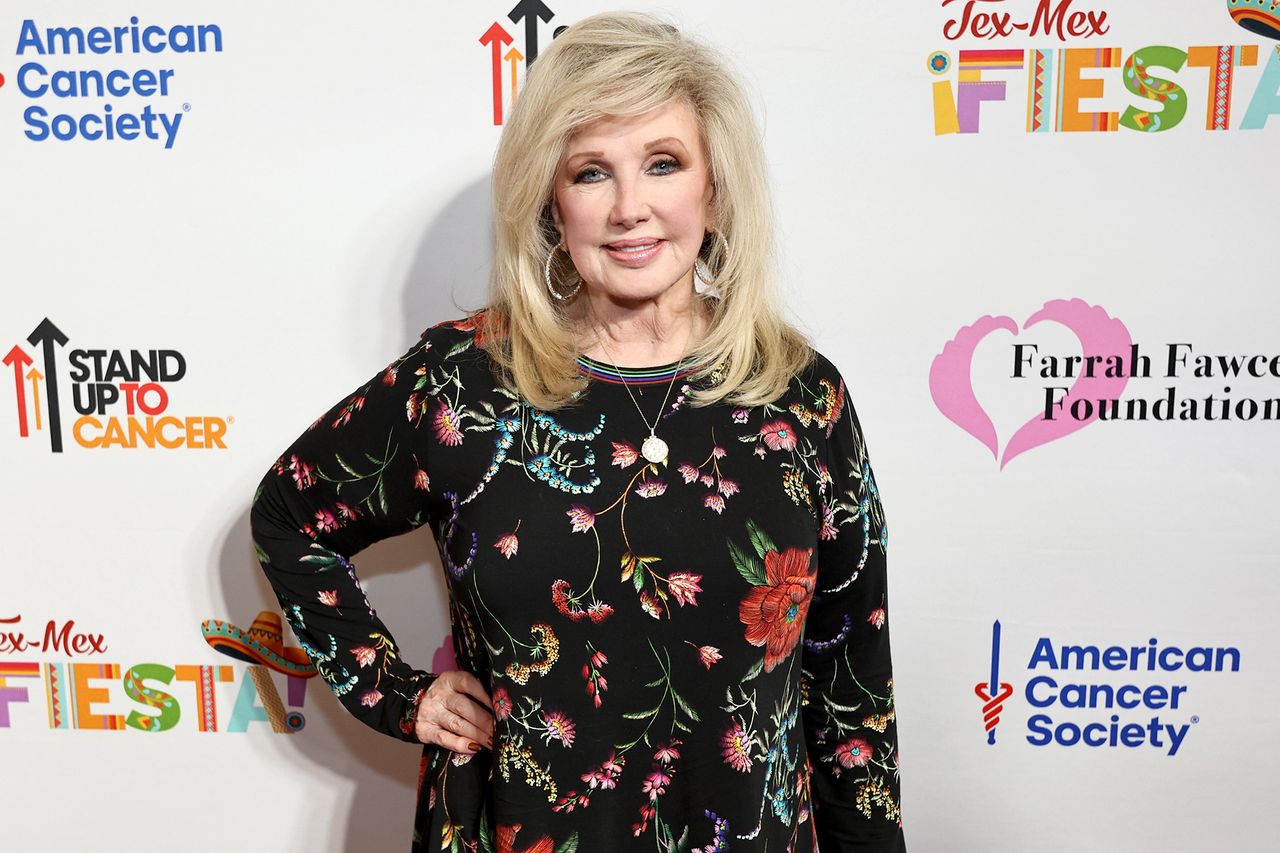 Morgan Fairchild at a charity event standing in front of a stepandrepeat banner with logos of sponsors