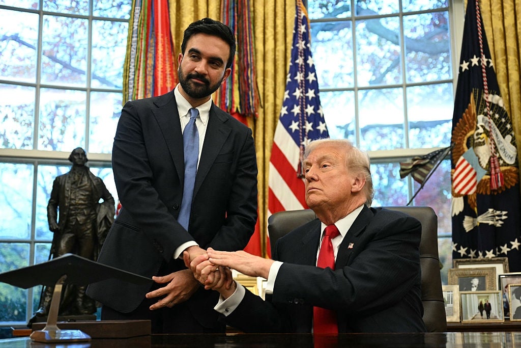 Two men are in an official-looking office. One is seated at a desk, reaching out to shake hands with the other, who is standing