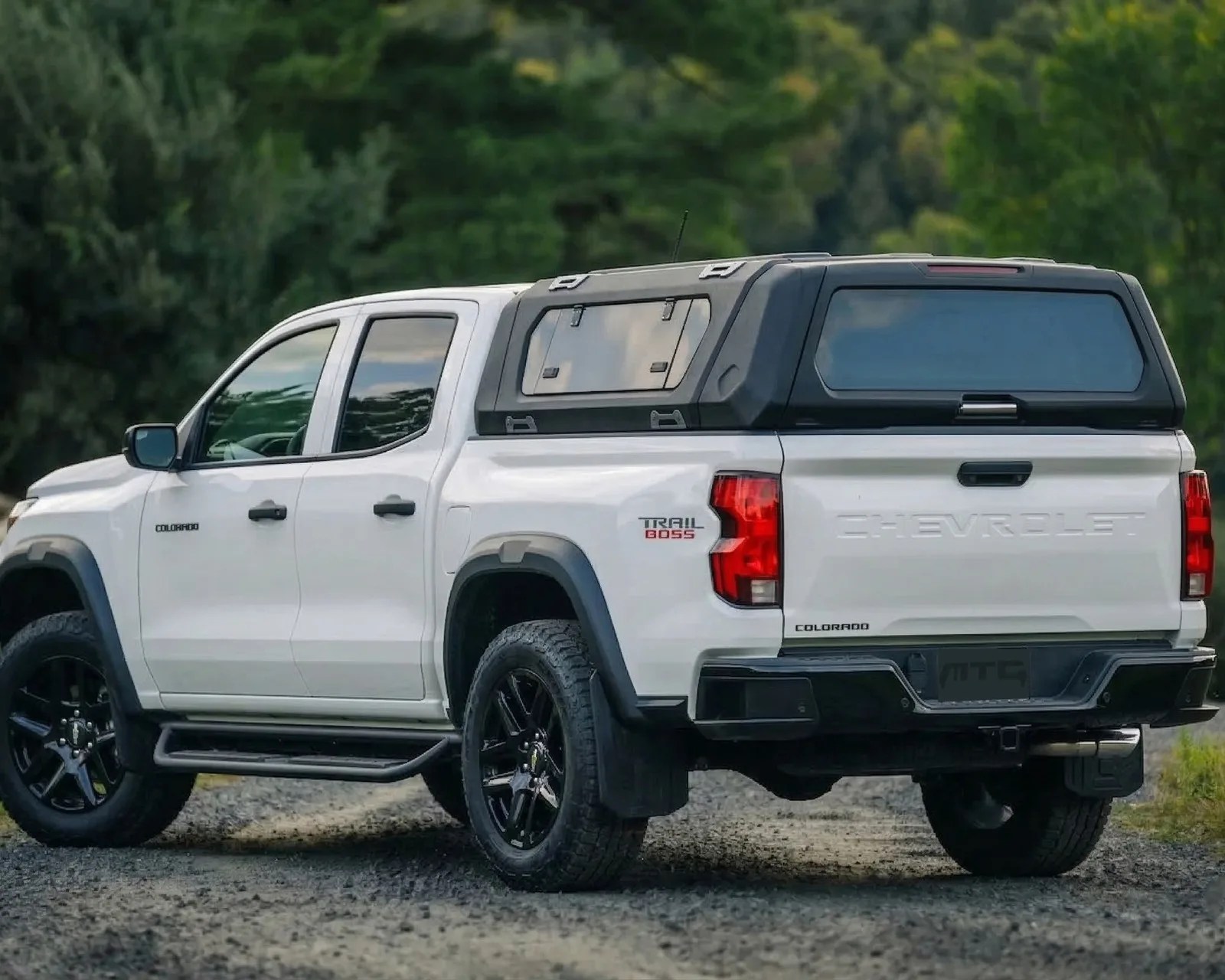White Chevrolet Colorado Trail Boss pickup truck with black camper shell and black wheels on gravel road.