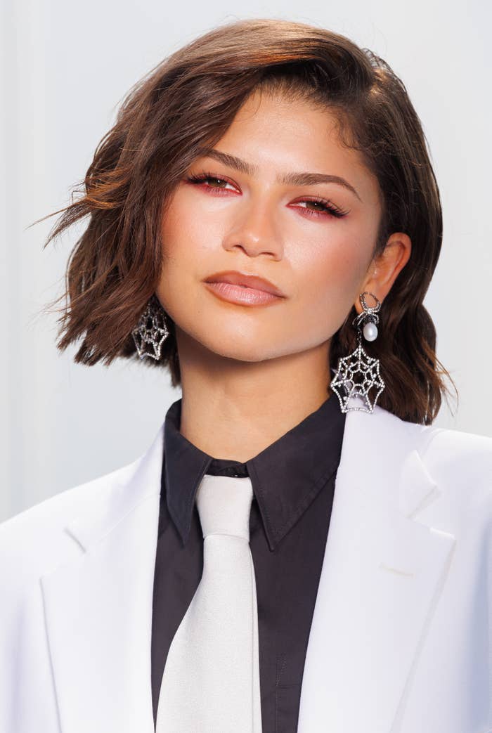 A person in a white suit, black shirt, and white tie, with stylized earrings, poses at an event