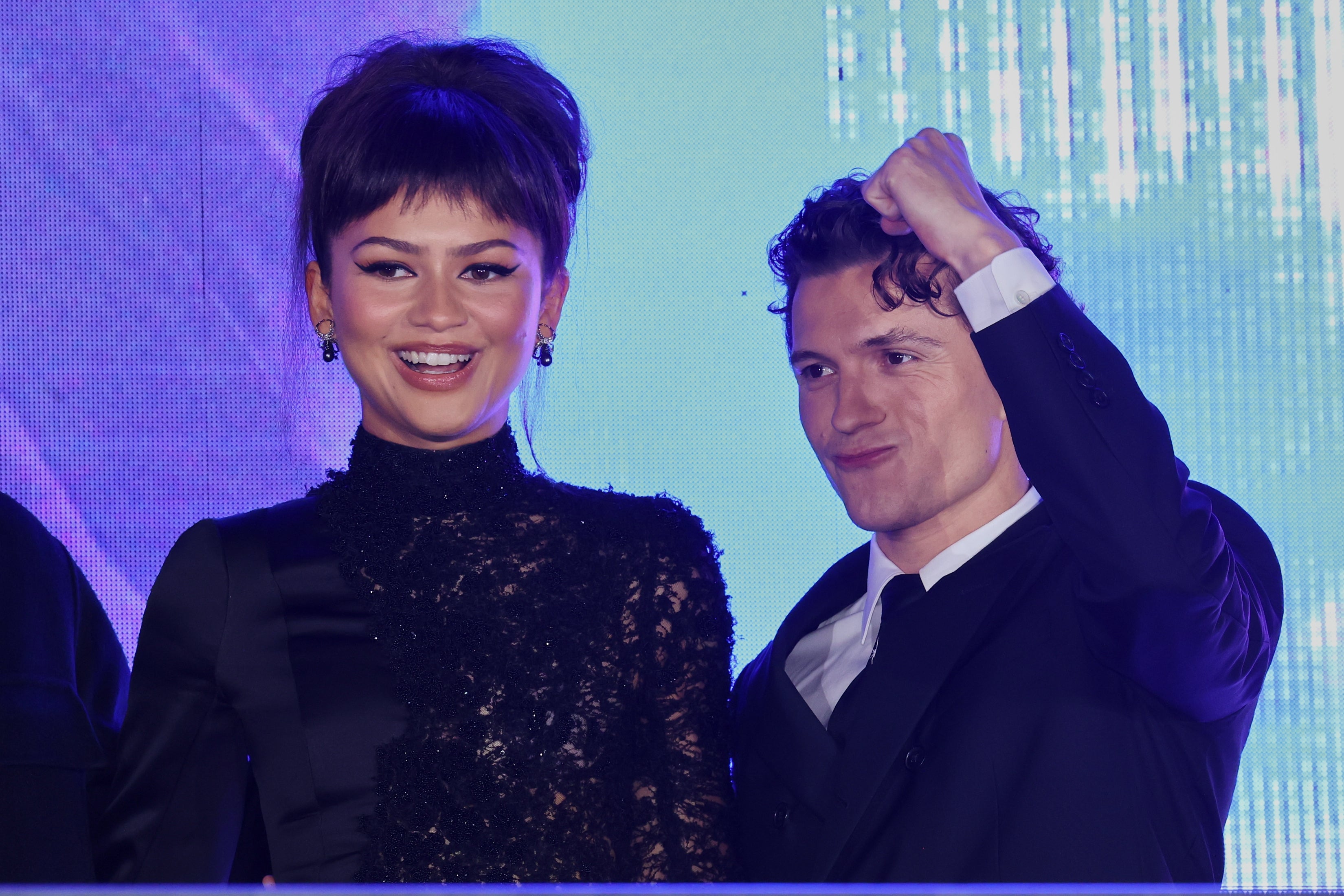 Two celebrities on stage; one wears a stylish lace dress while the other is in a classic suit, looking victorious and smiling