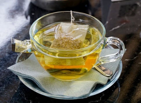 A cup of tea with a teabag in clear glass on a saucer with a spoon, resting on a napkin on a dark surface