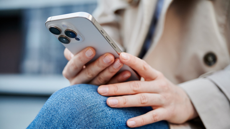 A closeup of an individual holding a white iPhone in their right hand as their left hand wrests on their knee, the image is cropped so that the iPhone is more prominent than the individual