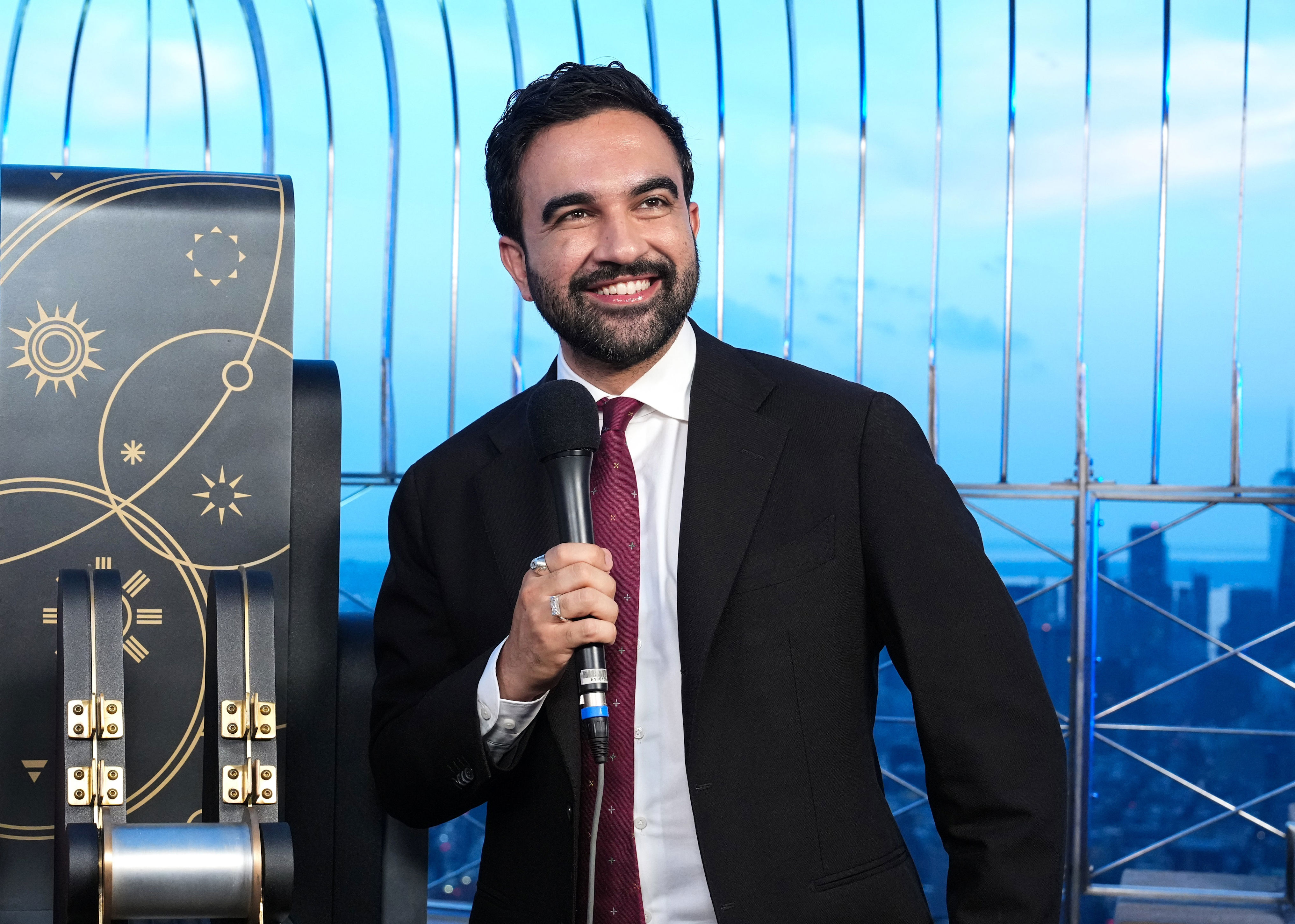 A man in a suit holds a microphone, smiling, with a cityscape backdrop and ornate decor beside him