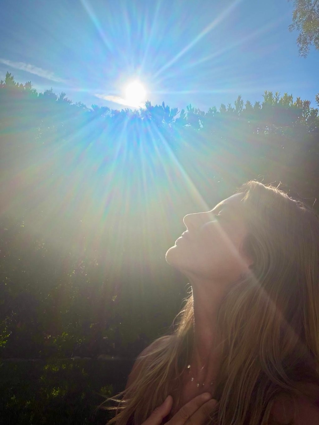 Gisele Bundchen looking up at the sun with a hand on her chest.