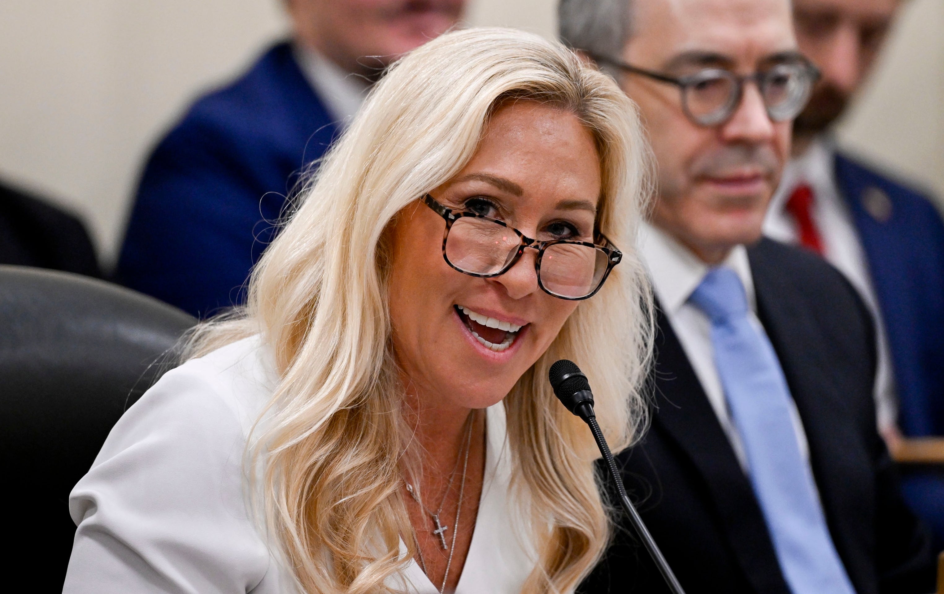 Person with glasses speaks into a microphone at a formal meeting; others sit in the background listening attentively