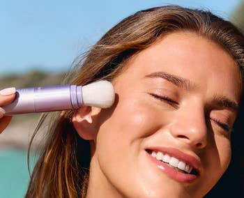 Person smiling while applying SPF 30 sunscreen with a brush on their face.