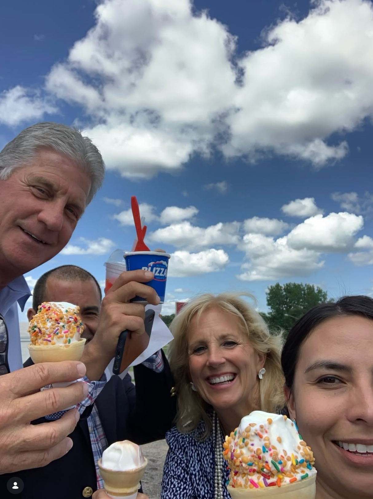 A group of people smiling outdoors, enjoying ice cream cones with sprinkles. A sunny day with scattered clouds in the background