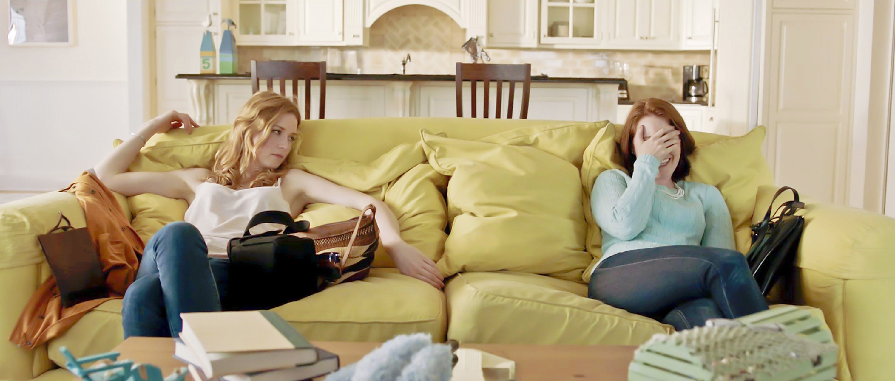 Two women sitting on a couch, one with a frustrated expression and the other covering her face with a hand, in a modern living room setting