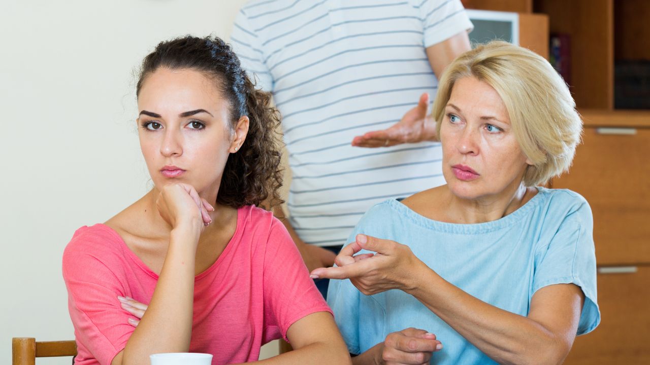 Two women sit at a table while a man stands behind them speaking.