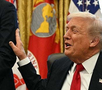 Two men in a formal setting, one seated with an expressive gesture, the other standing and smiling, flags displayed in the background