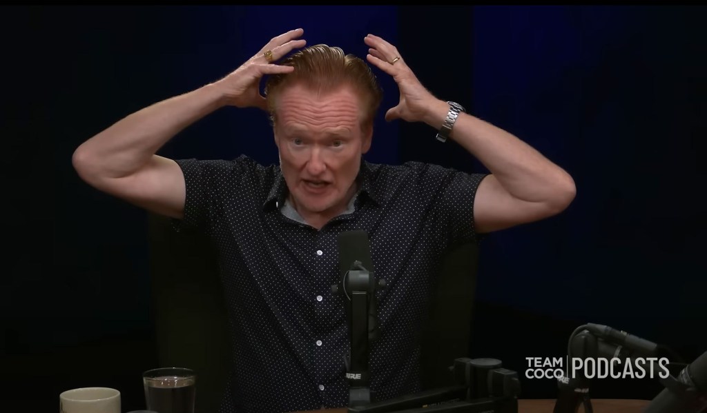 Conan O'Brien with his hands on his head, talking into a microphone.