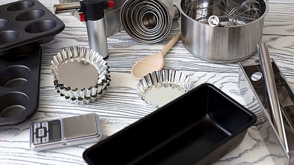 Various baking tools and utensils, including muffin tins, baking pans, metal molds, a kitchen scale, a saucepan with whisks, a wooden spoon, and measuring cups, are arranged on a patterned surface.