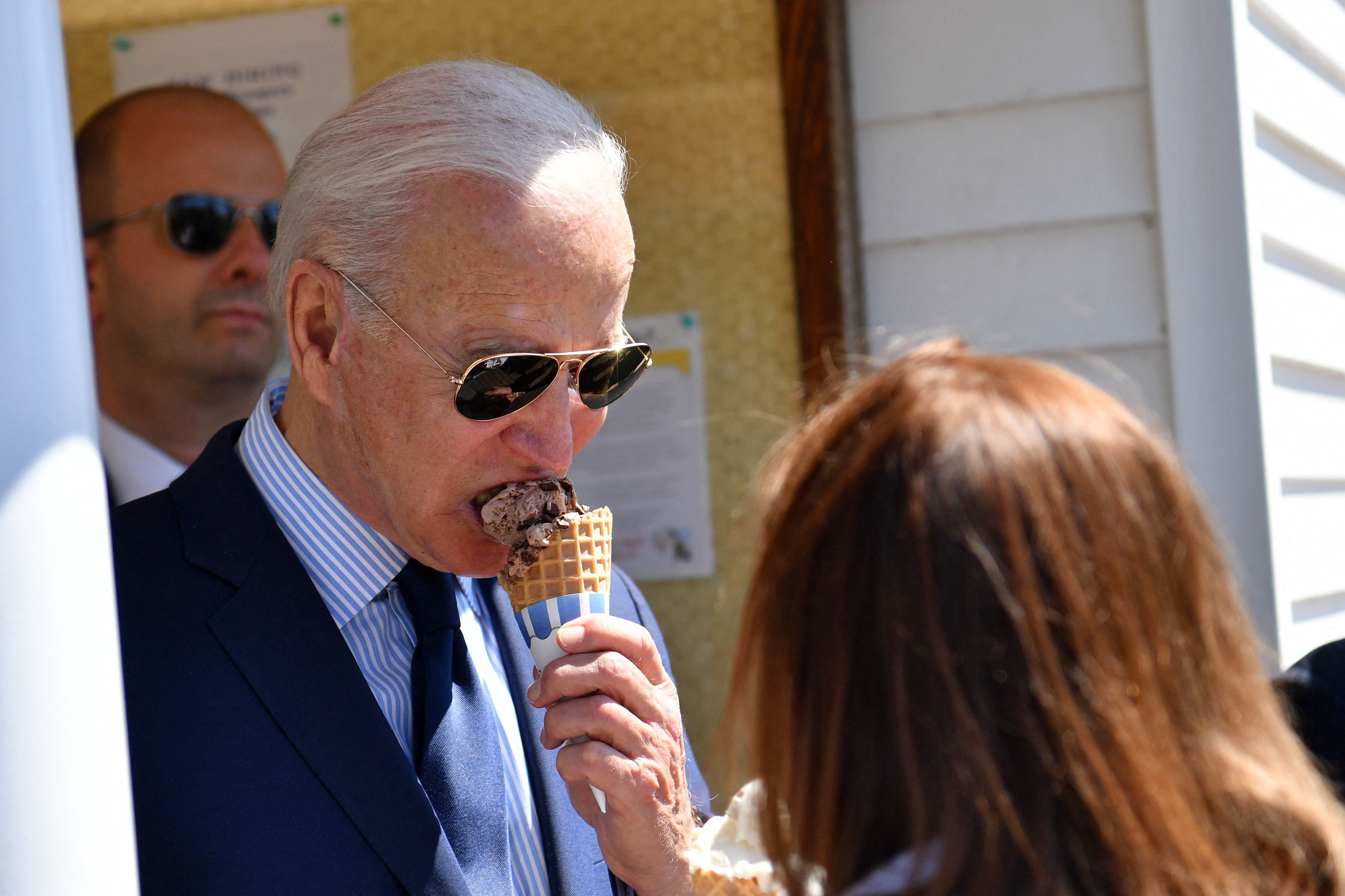 A person wearing aviator sunglasses enjoys an ice cream cone outside, with another person partially visible in the foreground