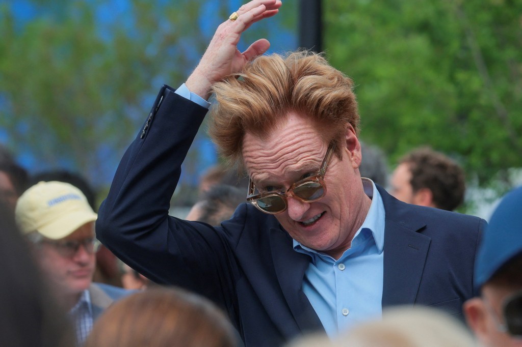 Conan O'Brien adjusting his red hair and smiling, wearing a blue shirt and suit jacket, with sunglasses on his nose.
