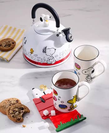 Snoopy-themed kitchen set featuring a kettle, two mugs with Snoopy and Woodstock designs, a Snoopy doghouse coaster, cookies, and sugar cubes