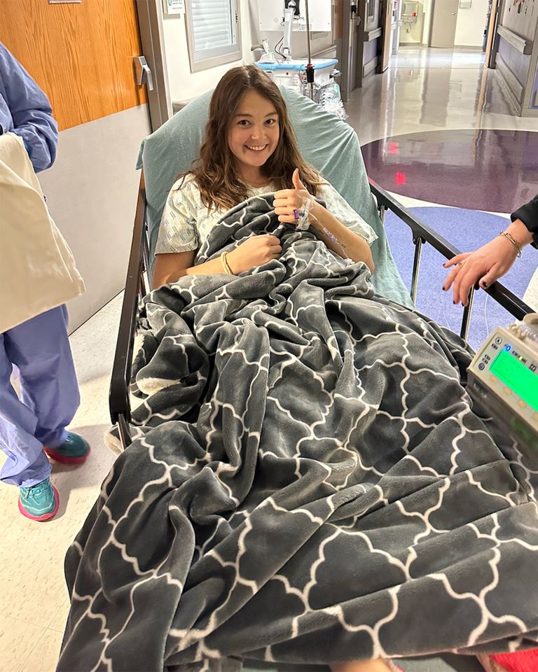 A woman in a hospital bed giving a thumbsup gesture covered with a patterned blanket while in a clinical setting