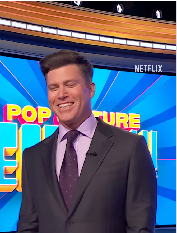 Man in a suit with a dotted tie smiling on a game show set with "Pop Culture" text in the background