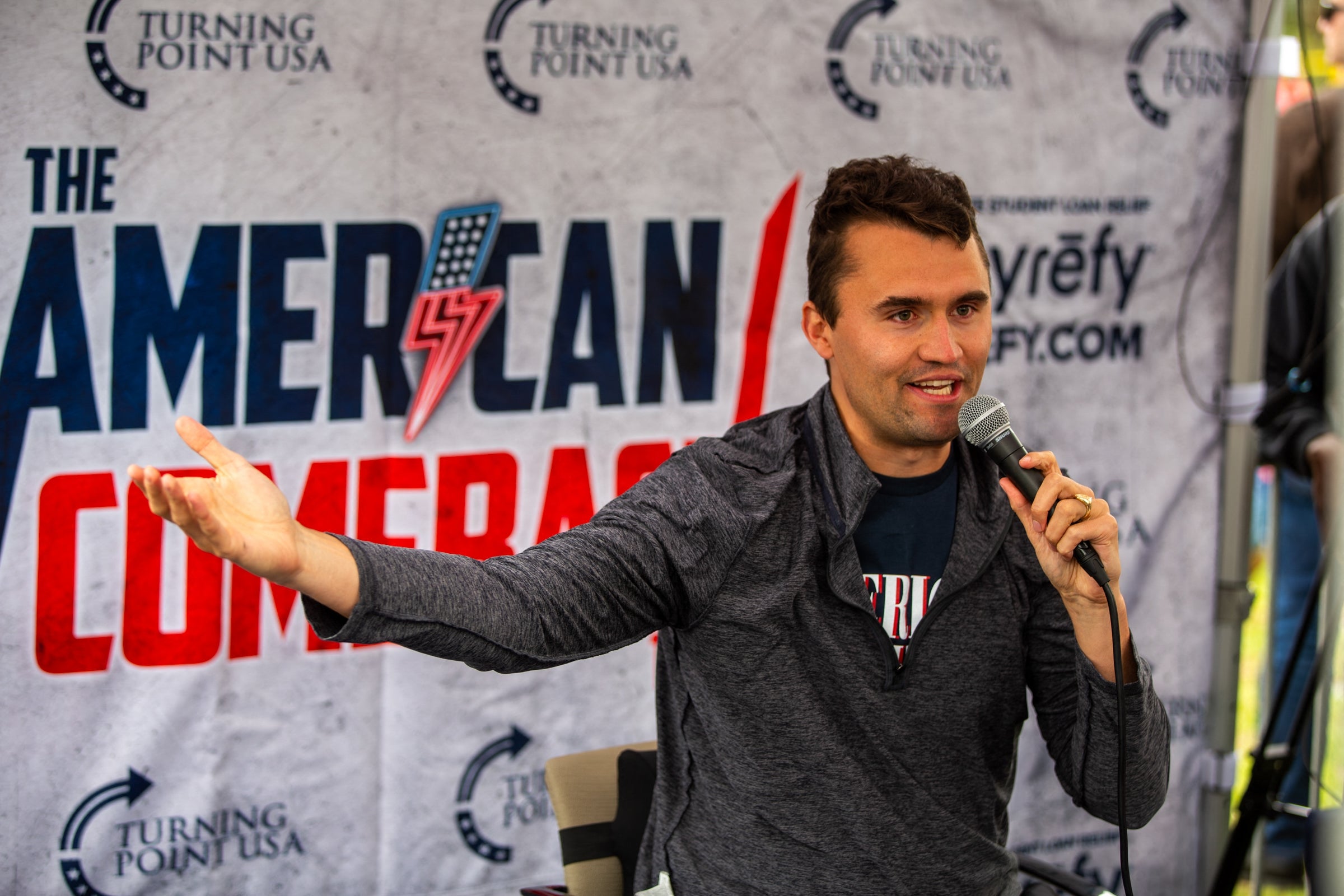 A person speaks into a microphone at an event with an "American" themed backdrop