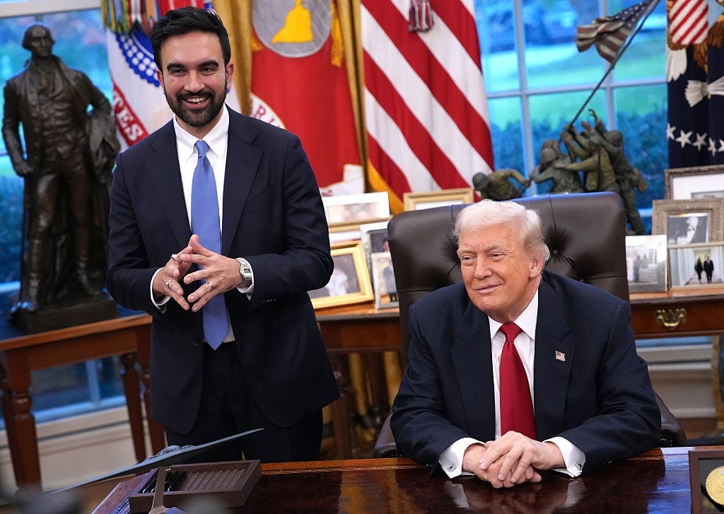 Two individuals are in an official setting. One is seated in a suit and red tie, while another stands next to him in a suit and blue tie, appearing cheerful