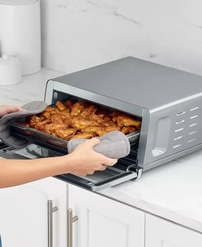 Person using oven mitts to remove a tray of cooked chicken wings from a countertop toaster oven in a kitchen
