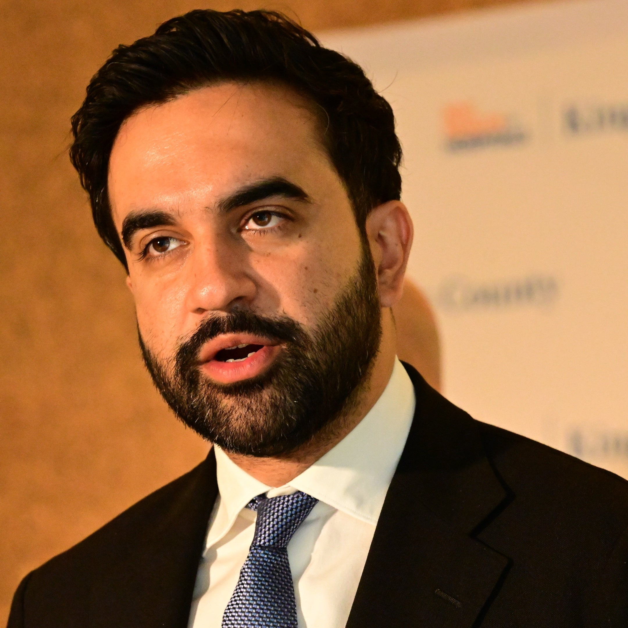 A person speaking at an event, wearing a suit and tie, with a backdrop displaying