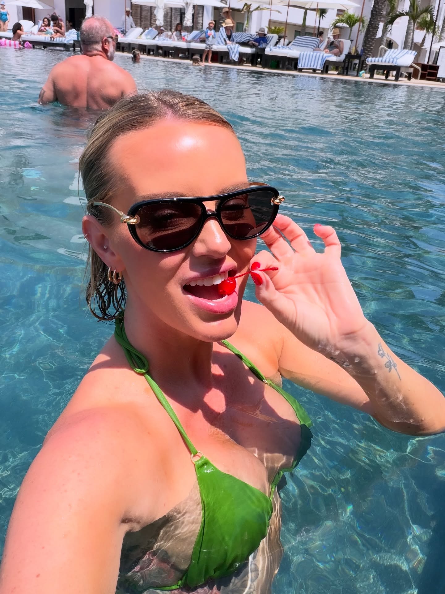 Whitney Rose in a green bikini with a cherry in her mouth, in a swimming pool.