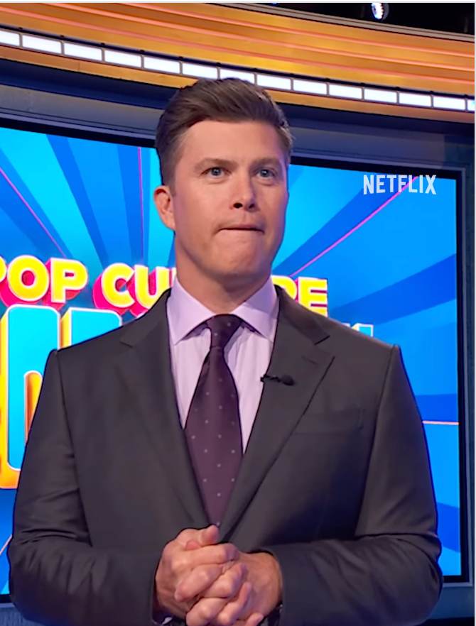 Person in a suit on a TV game show set, looking thoughtful, with a blue and yellow background