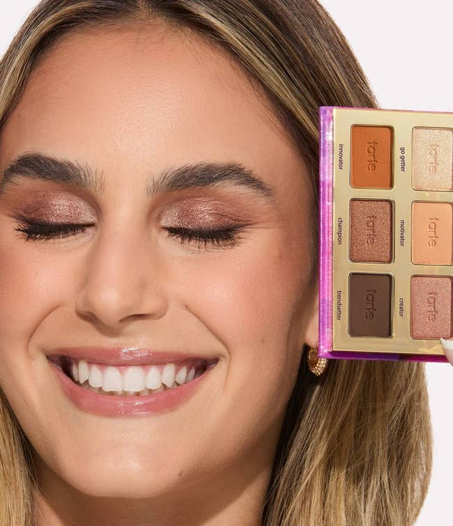 Person smiling with eyes closed, holding a makeup palette near face, showing eyeshadow shades
