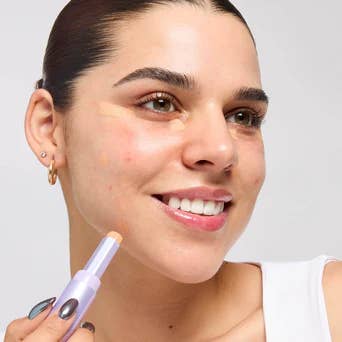 Model applying concealer stick on her face, smiling, with close-cropped hair and a white top