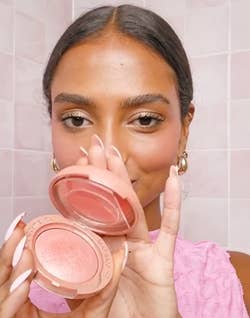 A person holds an open compact blush, smiling. They wear a textured top and gold hoop earrings