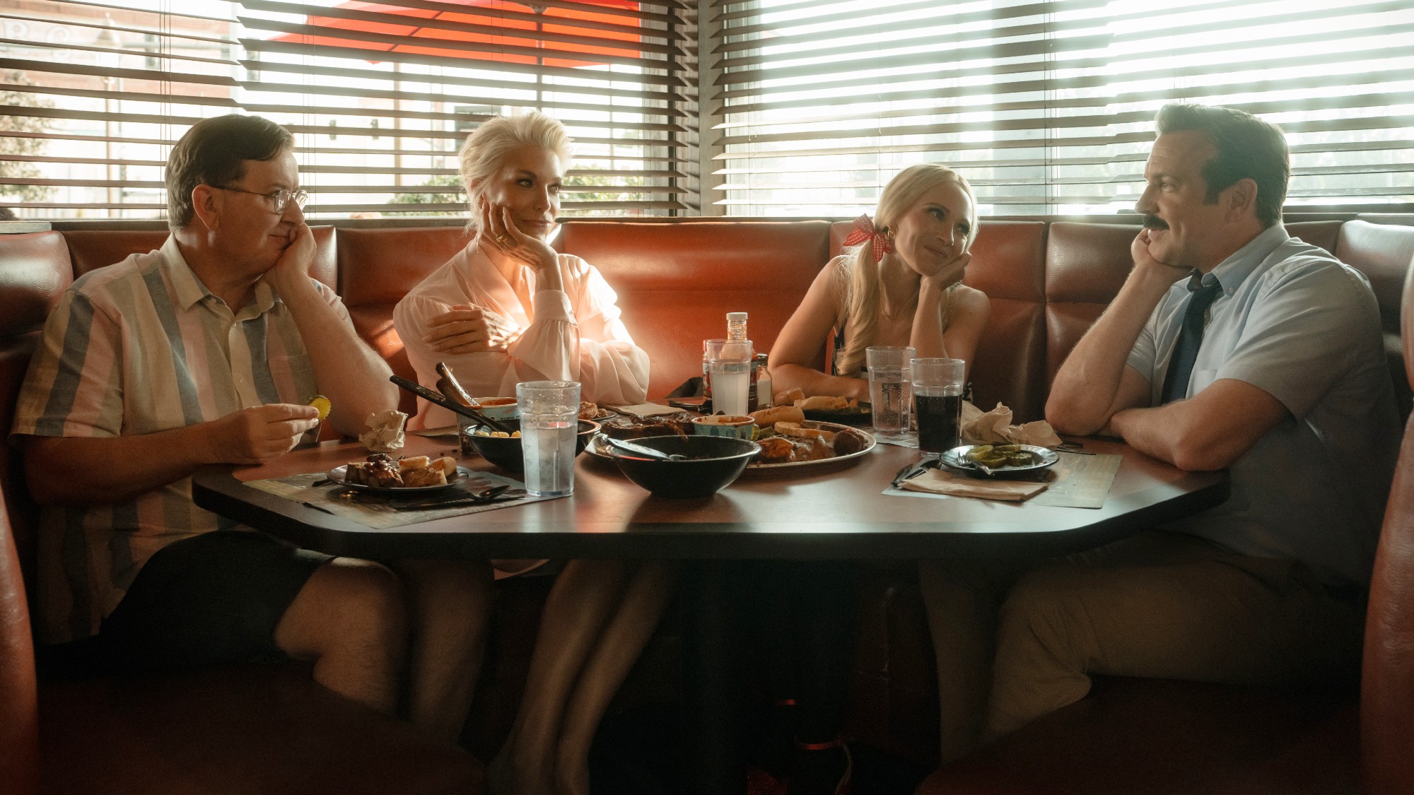 Jeremy Swift, Hannah Waddingham, Juno Temple and Jason Sudeikis all sitting together in a booth with one of their hands resting on their chins.