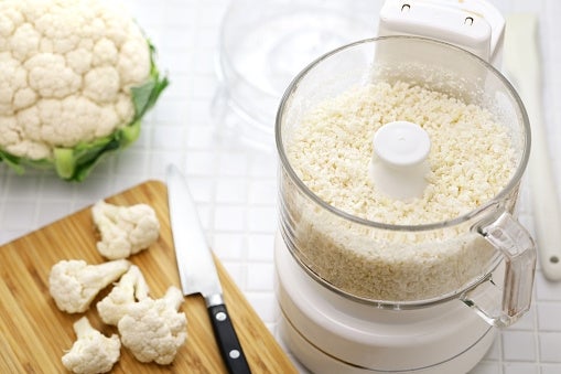 Cauliflower processed into rice-like texture in a food processor, with cauliflower florets and knife on a wooden cutting board nearby