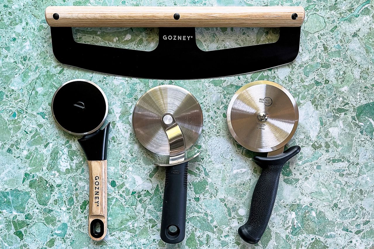 Four pizza cutters on a green surface including a large rocking blade and three rollingstyle cutters