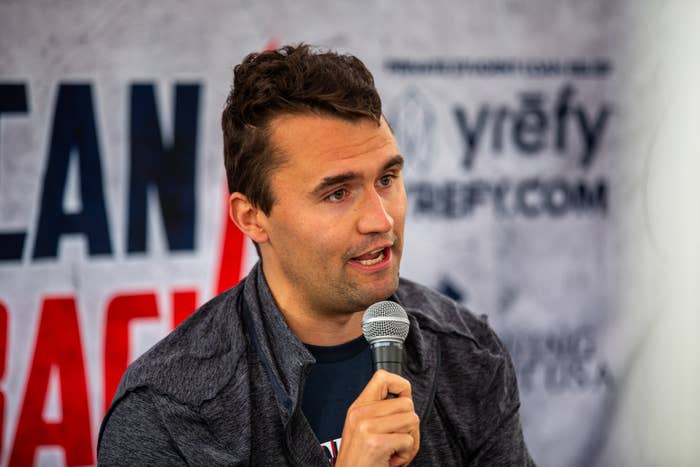 Person holding a microphone, speaking at an event with a branded backdrop