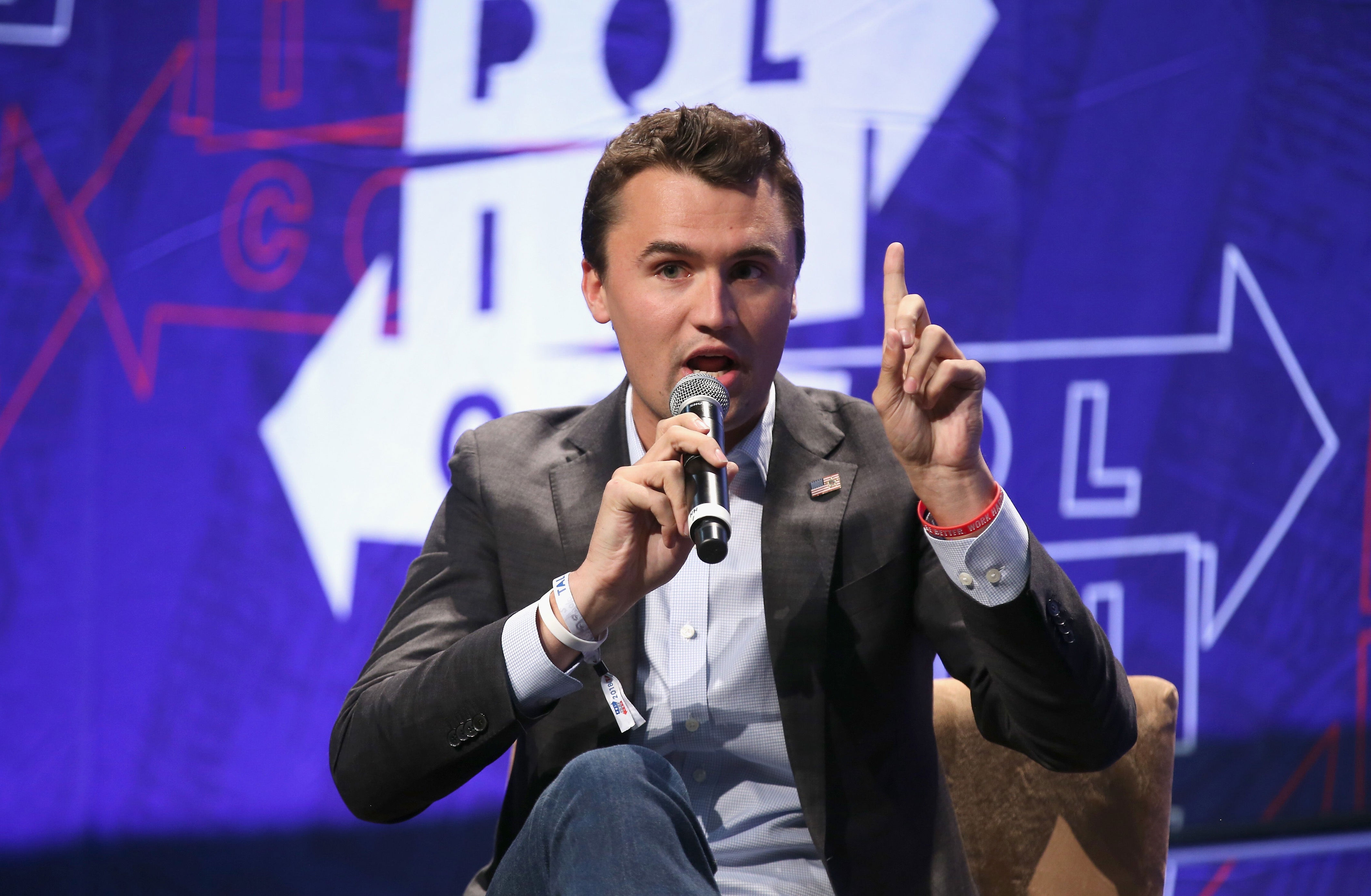 Person seated onstage speaking into a microphone, gesturing with one finger raised, wearing a blazer and jeans. Background shows directional arrows