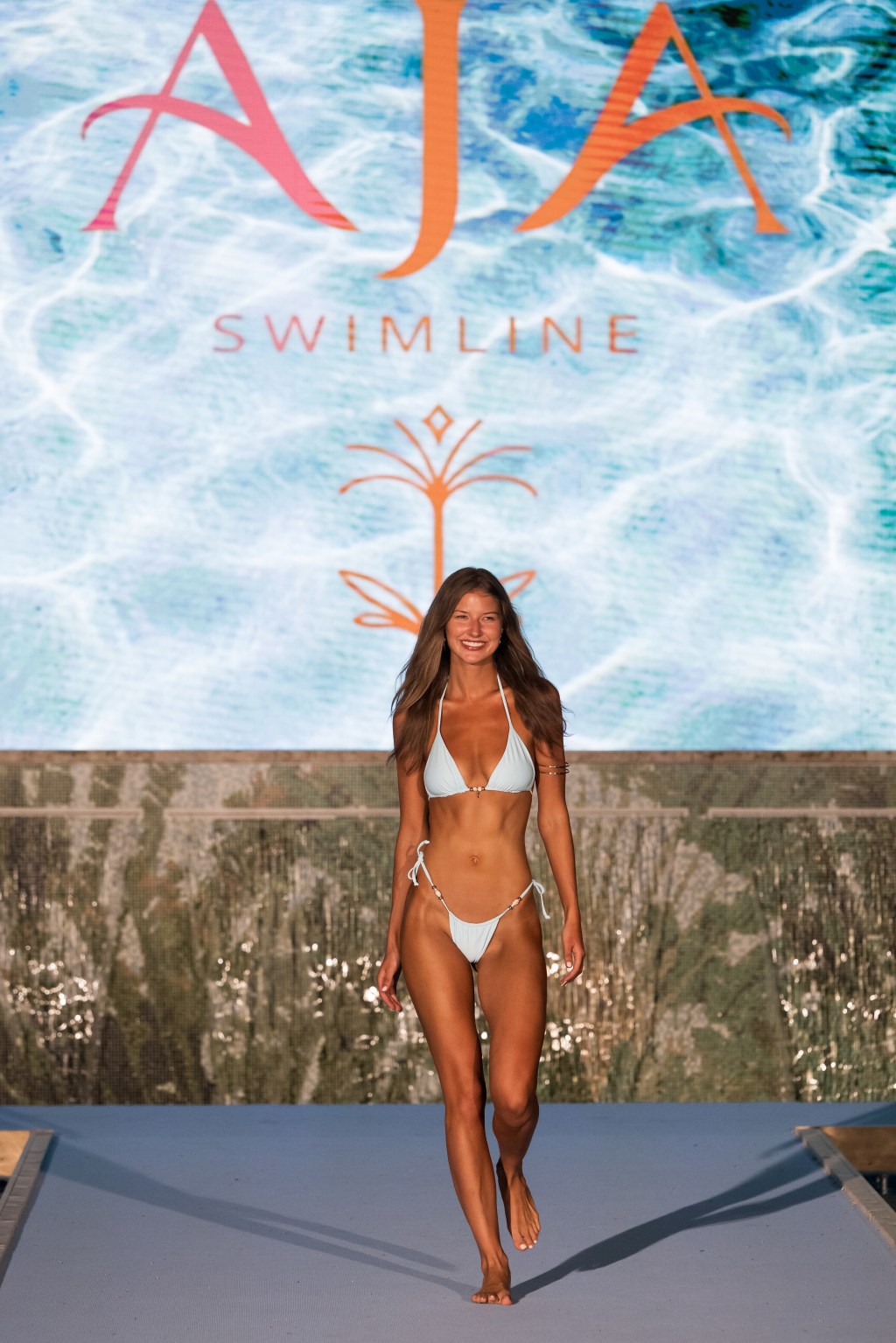 A model on a runway wearing a white Aja Swimwear bikini.